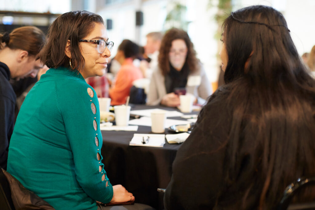 Two women sit at a table having a conversation during a group event, with other people and refreshments visible in the background.