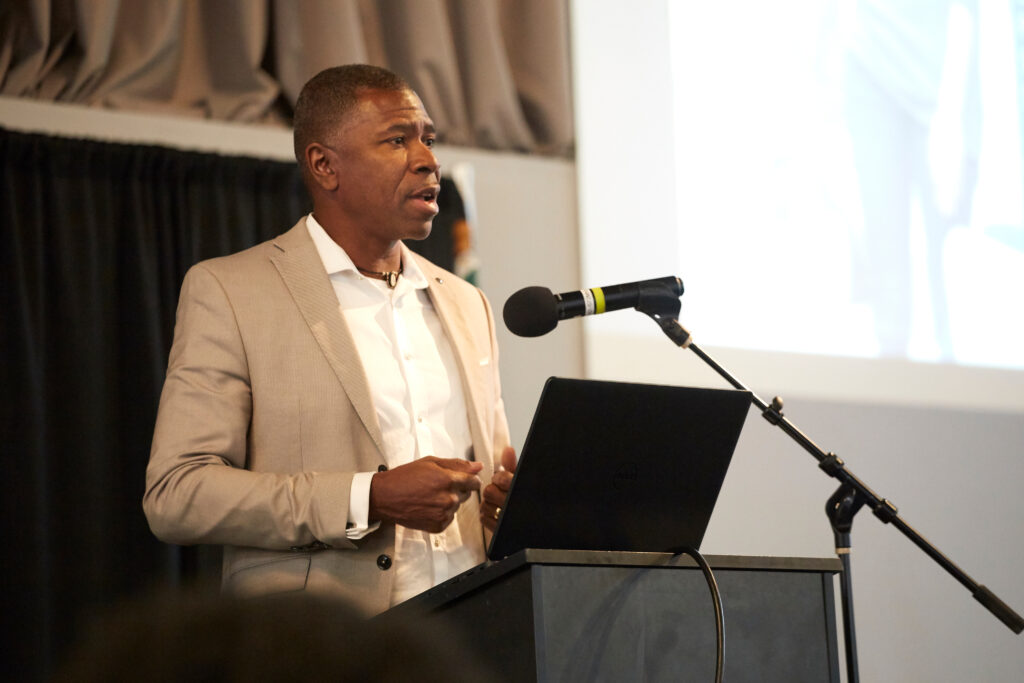 A man in a light-colored suit stands at a podium, speaking into a microphone with a laptop in front of him in a conference setting.