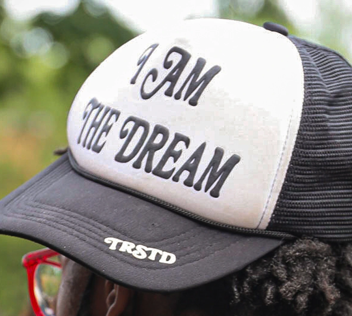 A person wearing a black and white trucker hat with the text "I AM THE DREAM" on the front and "TRSTD" on the brim.