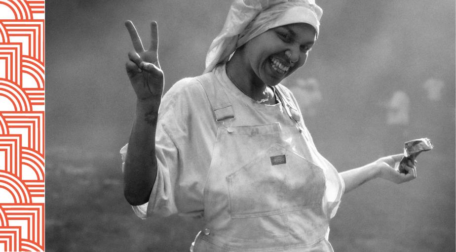 A person in white overalls and a headscarf smiles, holding a paintbrush in one hand and making a peace sign with the other.