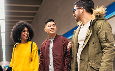 Three people walk together indoors, smiling and talking. One woman wears a yellow sweater, and two men wear jackets, one with a fur-lined hood.
