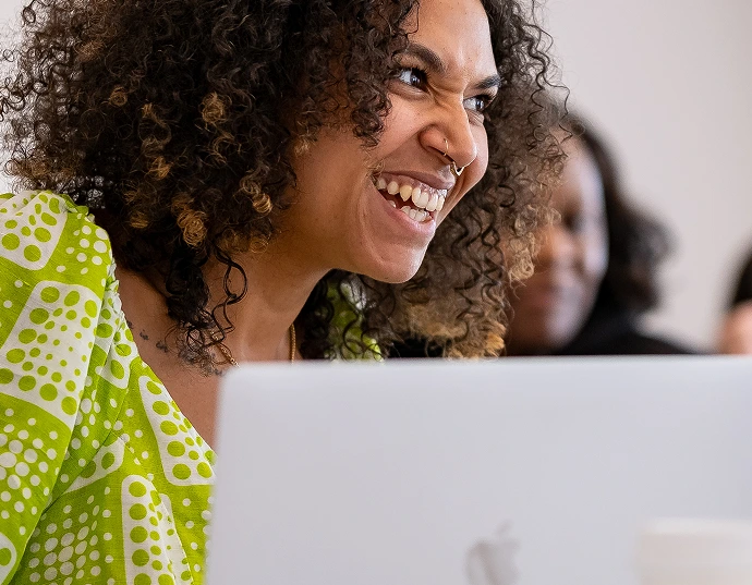 Person with curly hair and a nose ring smiling while sitting behind a MacBook laptop, wearing a green and white patterned top.
