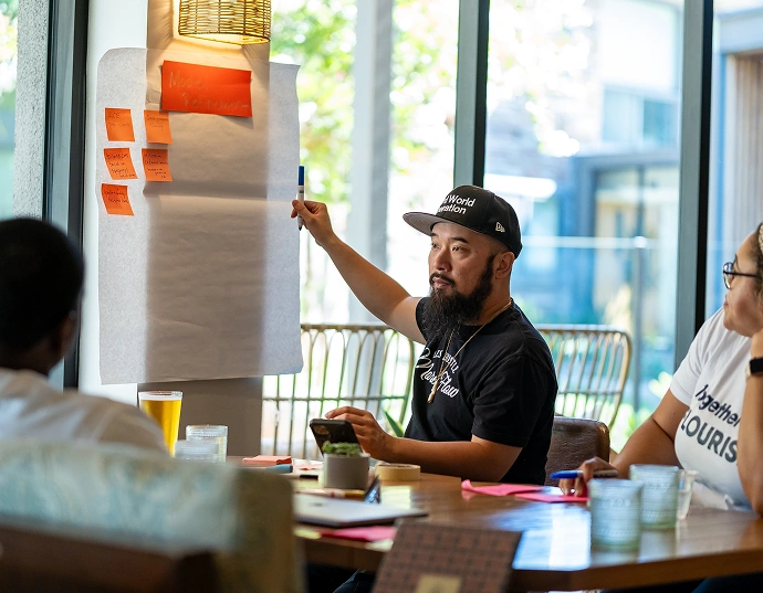 A man points to a blank sheet of paper with sticky notes on a wall while discussing with others seated at a table in a bright room.