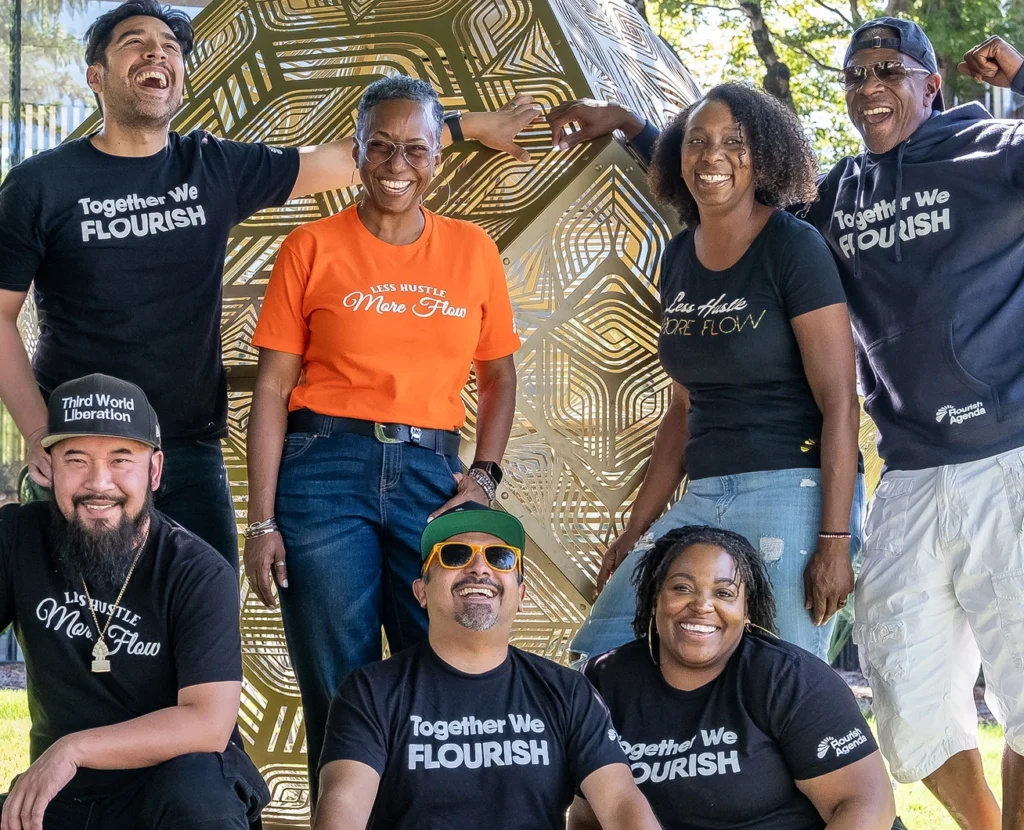 Seven people pose and smile outdoors in front of a large gold geometric sculpture, wearing t-shirts with motivational slogans such as "Less Hustle More Flow" and "Together We Flourish.