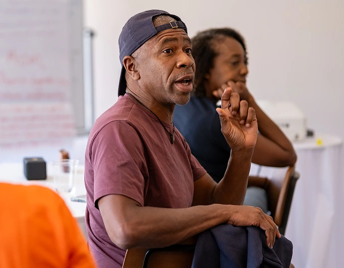 A man in a backward cap and maroon shirt gestures while speaking during a group discussion in a classroom setting. Another person sits in the background.
