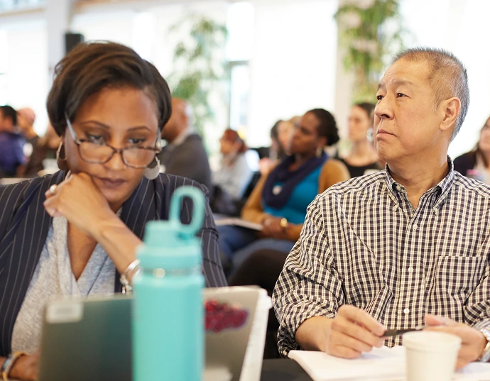 Two people sit at a table during a meeting or conference, one looking at a laptop and the other gazing forward, with notebooks and drinks in front of them.