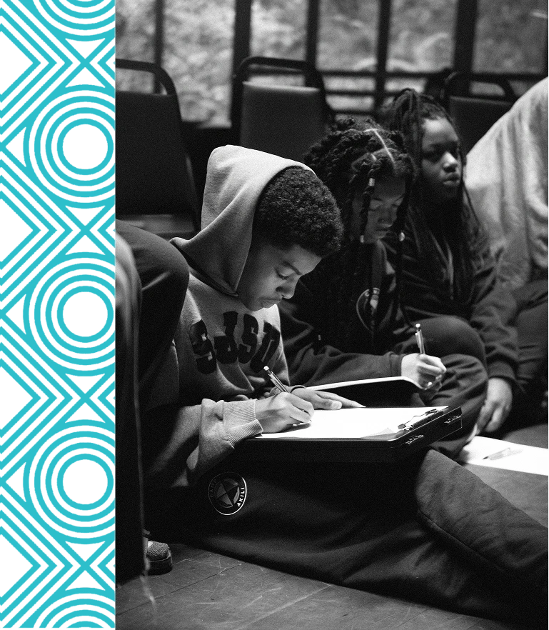 A group of young people sit on the floor, focused on writing in notebooks, with empty chairs and a window in the background.