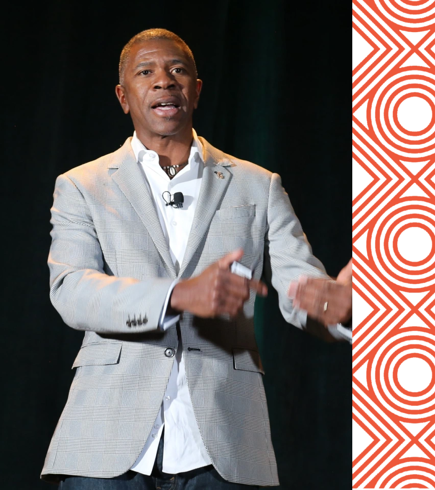 A man in a light gray blazer and white shirt is speaking on stage, gesturing with his hands, with a microphone clipped to his shirt.