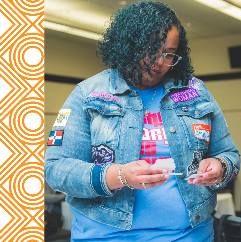 A woman with curly hair and glasses, wearing a denim jacket with colorful patches, looks down at an object in her hands in an indoor setting.