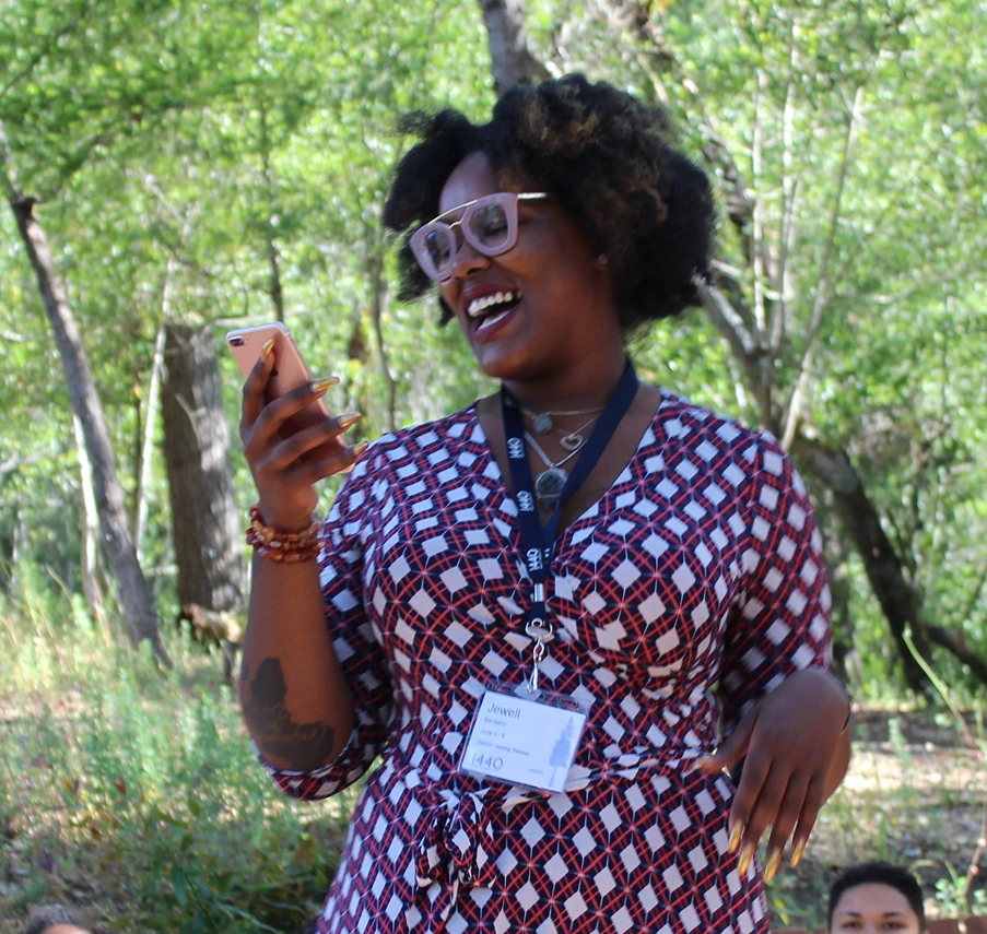 A woman with glasses and a patterned dress smiles while looking at her phone, standing outdoors with green trees in the background.