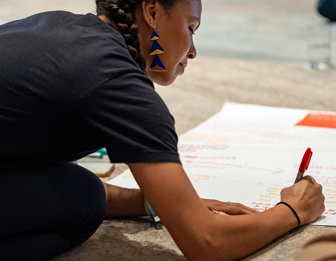A person with braided hair writes on a large sheet of paper with a red marker while kneeling on the floor.