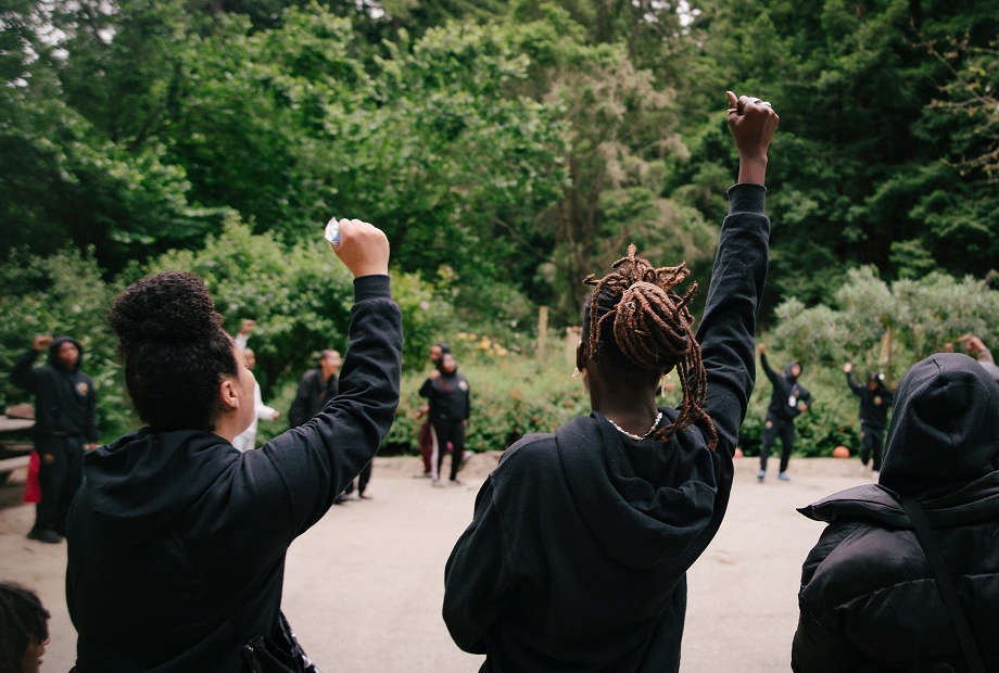A group of people gathered outdoors, with two individuals in the foreground raising their fists in the air. Lush green trees and bushes surround the group.