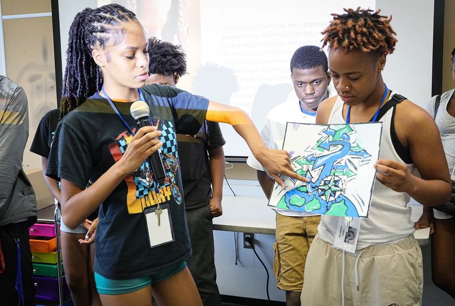 A young person holds a microphone and points to a drawing held by another person during a group presentation in a classroom.