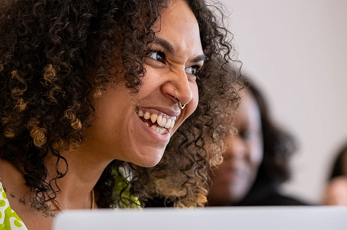 A person with curly hair and a nose ring smiles widely, showing their teeth, while sitting in front of a blurred background with another person.