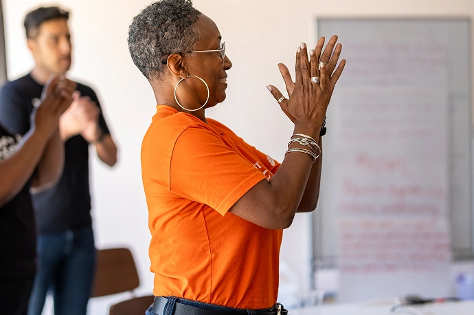 A woman in an orange shirt stands clapping, with a whiteboard and other people visible in the background.