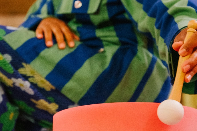 A person wearing a blue and green striped garment holds a mallet and strikes a pink drum or bowl.