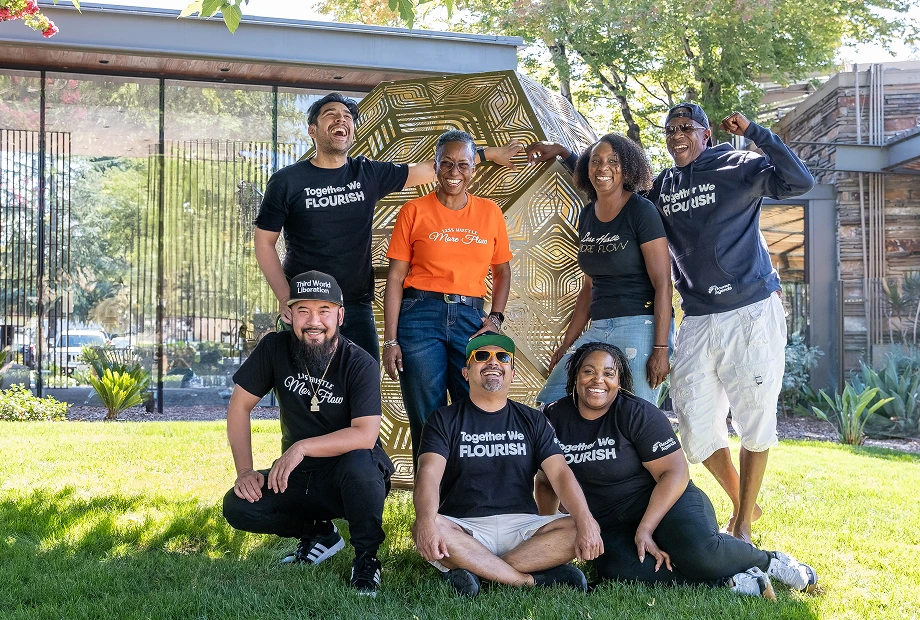 A group of seven adults poses and smiles on a lawn in front of a large golden geometric sculpture and modern glass building.