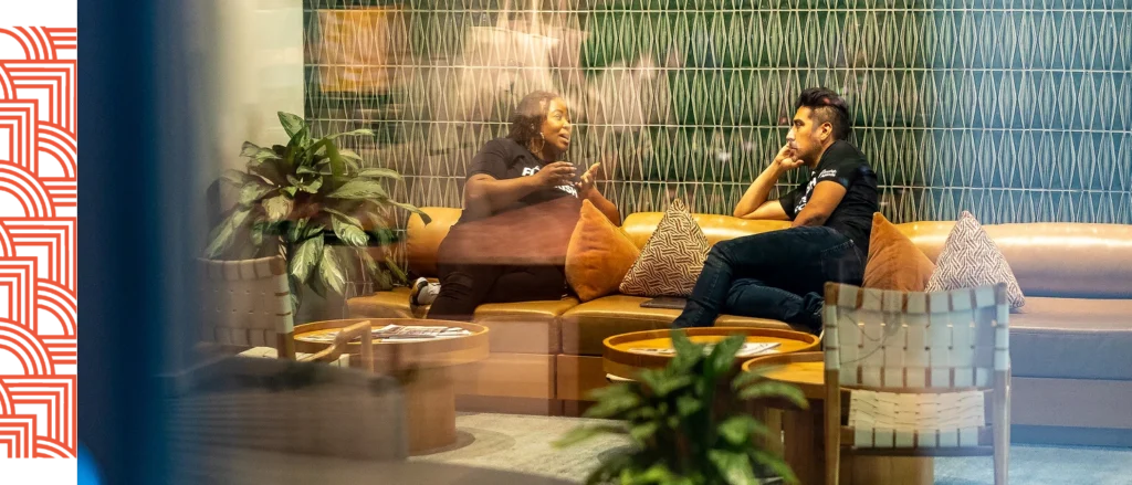 Two people sit on a curved sofa in a modern lounge, engaged in conversation. Decorative pillows and indoor plants are on the coffee table and surrounding area.