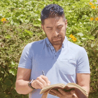 A man in a light blue polo shirt stands outside, writing in a notebook with a pen, surrounded by greenery and yellow flowers.