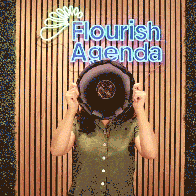 A person in a green shirt holds a speaker in front of their face, standing in front of a wall with a neon "Flourish Agenda" sign.