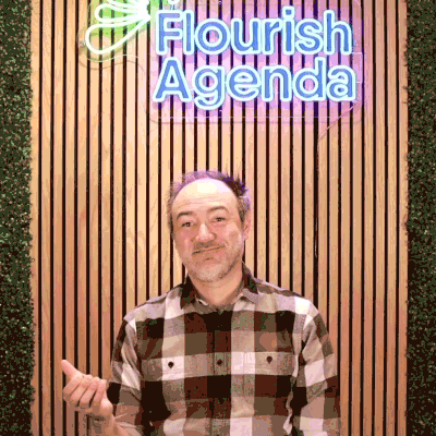 A person in a plaid shirt gestures with one hand while standing in front of a wall with a neon sign that reads "Flourish Agenda.