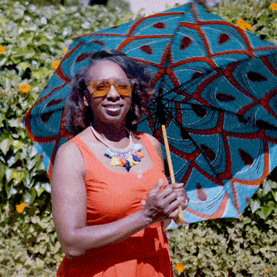 A woman in an orange dress and sunglasses stands outdoors holding a colorful, patterned umbrella. Green foliage and yellow flowers are in the background.