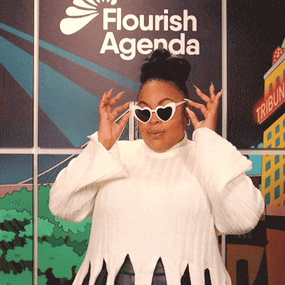 A person in a white sweater puts on heart-shaped sunglasses and poses in front of a backdrop with the words "Flourish Agenda.