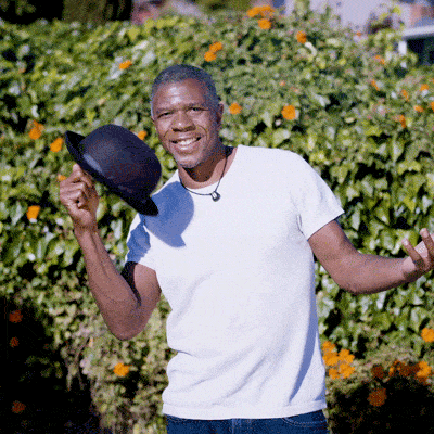 A man in a white t-shirt stands outdoors, smiling, and tipping a black hat with one hand. There are green bushes and orange flowers in the background.