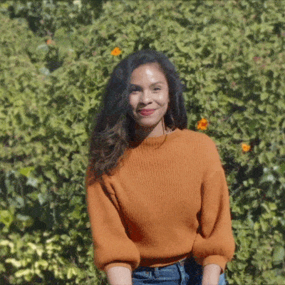 A woman wearing an orange sweater stands outside in front of green foliage, smiling and waving at the camera.