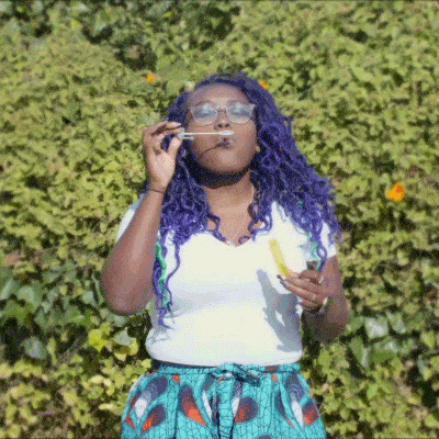 A person with purple hair and glasses blows soap bubbles outdoors, standing in front of a leafy green background.