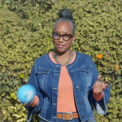 A woman in a denim jacket and glasses tosses a small blue ball while standing outdoors in front of green foliage.